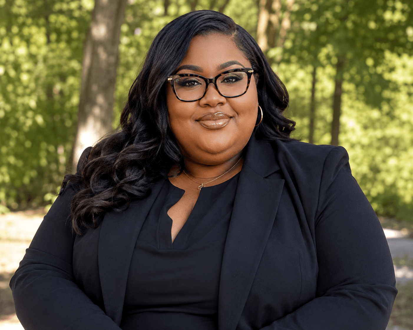 Smiling Black woman with long wavy hair, wearing glasses and a black blazer outdoors.
